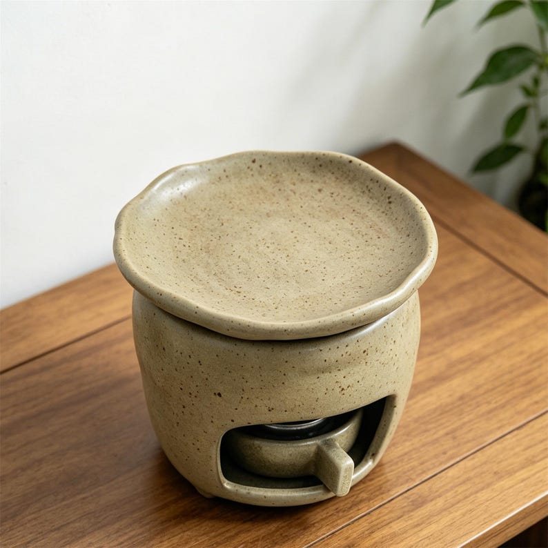May include: A beige ceramic essential oil warmer with a speckled finish. The warmer has a round base with a small opening and a removable dish on top. The design is simple and modern, suitable for aromatherapy and home fragrance.
