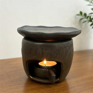 May include: A dark brown ceramic essential oil diffuser with a wavy-edged top and a tea light candle burning inside. The diffuser has a rounded body with a small handle and a textured surface. The candle flame glows brightly.