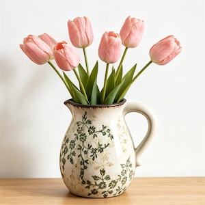 Peut inclure: Une cruche en céramique avec une finition émaillée craquelée de couleur crème et un motif de vigne florale vert foncé. La cruche présente une anse incurvée et un bec verseur, et elle est remplie de sept tulipes rose tendre sur tiges vertes.