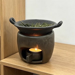 May include: A dark gray ceramic essential oil diffuser with a small dish containing dried herbs. A lit tea light candle sits inside the base, illuminating the interior. The diffuser has a rustic, handcrafted appearance.