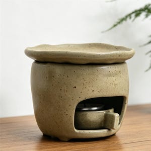 May include: A beige ceramic essential oil diffuser with a speckled finish. The diffuser has a round base with a small opening for a tea light and a separate, wavy-edged dish on top. The design is simple and natural.