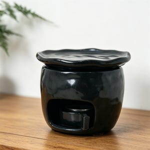 May include: A black ceramic essential oil warmer with a glossy finish. The warmer has a round base, a small opening, and a wavy-edged dish on top. It sits on a wooden surface.