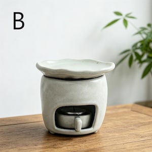 May include: A light gray ceramic essential oil diffuser with a wavy-edged dish on top. The diffuser has a rectangular opening to hold a small candle. The letter "B" is in the upper left corner. The diffuser sits on a wooden surface.