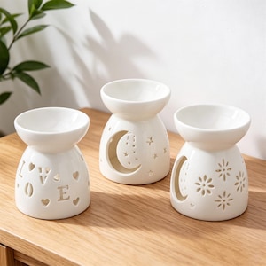 May include: Three white ceramic oil diffusers with decorative cut-out designs. One diffuser has the word "LOVE" cut out, another features a crescent moon and stars, and the third has flower shapes. Each has a bowl-shaped top.