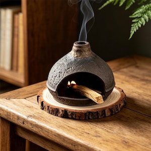 May include: A dark gray ceramic incense burner with a textured surface, burning a stick of palo santo. Smoke rises from the top. The burner sits on a wooden slice, placed on a wooden table. The background includes a bookshelf and greenery.