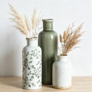 Puede incluir: Tres jarrones de cerámica con acabado de esmalte craquelado. Un jarrón alto y esbelto es de color verde salvia sólido. Dos jarrones más cortos son blancos, uno con un patrón floral verde y el otro con acabado craquelado y borde marrón moteado.