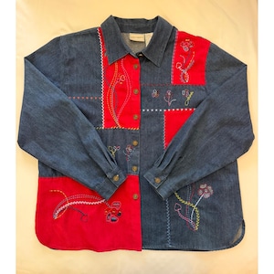 May include: A long-sleeved button-down shirt made of blue denim and red fabric panels. The garment features decorative multi-colored floral embroidery and cross-stitch detailing across the front. It has a standard collar and buttoned cuffs.