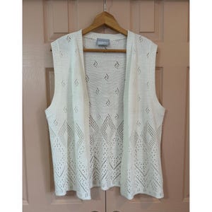 May include: A sleeveless, open-front vest in an off-white color, featuring a knit construction with a repeating diamond-shaped eyelet pattern throughout. The garment includes a ribbed trim along the armholes and front edges, with a Blair brand label.