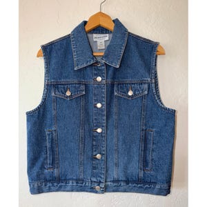 May include: A medium blue denim vest featuring a classic collar, a front button-down closure with silver-toned metal buttons, two chest pockets with buttoned flaps, and two vertical side pockets. A Pendleton brand label is visible at the neck.