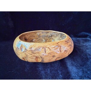 May include: A tan, oval-shaped ceramic bowl with a textured, mottled brown finish. The exterior features a series of evenly spaced, shallow vertical indentations around the perimeter, while the interior shows a dark, uneven, and rustic glaze.