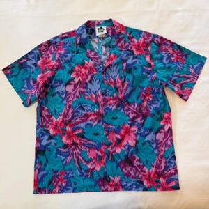 May include: Short-sleeved button-down shirt with a vibrant floral pattern in teal, pink, purple, and blue. The shirt has a classic collar and a relaxed fit, perfect for casual wear. The fabric appears to be lightweight and suitable for warm weather.