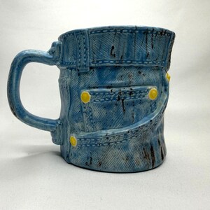 May include: A ceramic mug shaped like a pair of blue denim jeans, featuring a textured fabric pattern, a handle on the left side, and yellow painted details resembling metal rivets on the pockets and waistband, with dark brown distressed markings.