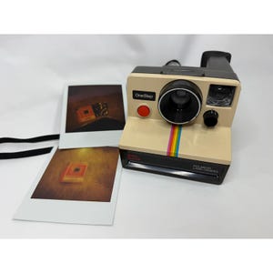 May include: A beige and black Polaroid OneStep Land Camera featuring a prominent red shutter button, a black lens, and a vertical rainbow stripe on the front. Two square instant photographs with dark, warm-toned imagery are placed beside the camera.