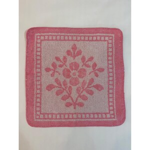 May include: A square pink and white cotton washcloth featuring a central floral motif with leaves and a flower, surrounded by a decorative border of small squares. The fabric has a textured terry cloth finish with a consistent color pattern.