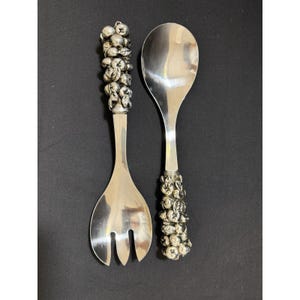 May include: A silver salad serving set with ornate handles. The fork has three tines, and the spoon has a large, oval bowl. The handles are decorated with a cluster of small, round, silver-colored shapes, possibly berries or grapes.