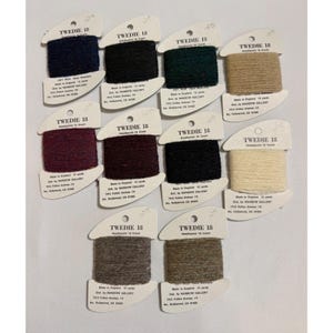 May include: A collection of spools of Tweedy 18 yarn in various colors, including navy, black, green, tan, burgundy, and cream. Each spool is labeled with the brand name and details. The yarn is made of 100% wool.
