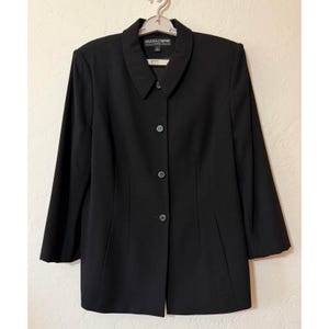 May include: A solid black blazer featuring a pointed collar, long sleeves, and a front button closure with four visible buttons. The garment includes vertical seam detailing along the front panels and a label reading Kasper & Company A.S.L.
