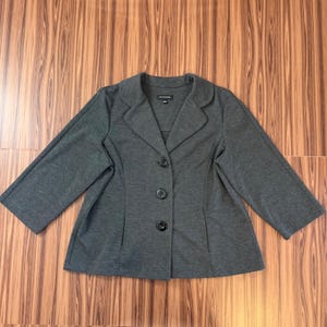 May include: A gray blazer with three black buttons and a notched collar. The blazer has long sleeves and is made of a soft material. The blazer is laid flat on a wooden surface.