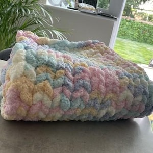 May include: A chunky hand-knitted blanket featuring a soft, variegated color palette of pastel pink, baby blue, creamy yellow, and white. The throw is crafted with thick, plush yarn in a consistent, textured loop stitch pattern, folded neatly.