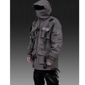 Puede incluir: Chaqueta t&aacute;ctica gris con m&uacute;ltiples bolsillos, correas y una capucha que cubre la cara. La chaqueta tiene el texto "H-G" en el pecho y el brazo. La chaqueta es parte de un atuendo futurista de estilo urbano.