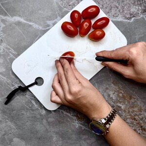 May include: A white cutting board with a marble effect, holding several red tomatoes. A person is slicing a tomato with a black-handled knife. The board has a black loop. The text "Daily Design" is visible. Tomatoes are being prepared.