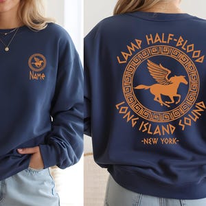 Op de afbeelding: Marineblauw sweatshirt met een oranje Pegasus-logo en de tekst "Name" op de voorkant. De achterkant toont de tekst "Camp Half-Blood Long Island Sound - New York" in oranje. Een casual kledingstuk met lange mouwen.