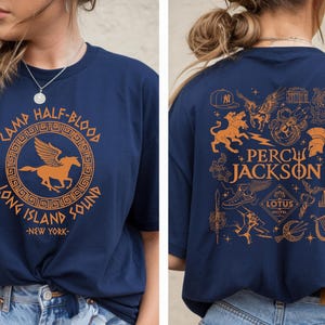 Puede incluir: Una camiseta azul marino con estampados gráficos en naranja. El frente muestra un logotipo circular de Camp Half-Blood Long Island Sound New York con un pegaso. La espalda presenta varios iconos temáticos de Percy Jackson y el texto Percy Jackson.
