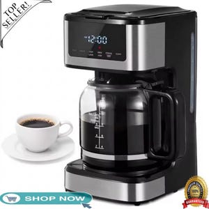 May include: Black and silver coffee maker with a glass carafe and digital display showing 12:00. A white cup of coffee sits on a saucer to the left. The carafe has measurements for 2, 4, 6, 8, 10, and 12 cups.