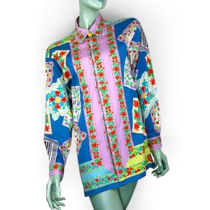 Könnte beinhalten: Ein langärmeliges Hemd mit einem lebendigen Blumen- und geometrischen Muster. Das Hemd hat einen blauen Hintergrund mit rosa und roten Blumenstreifen auf der Vorderseite. Die Ärmel und der Kragen haben eine Mischung aus Blumen- und geometrischen Designs.