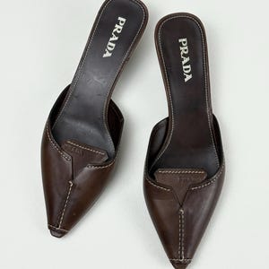 Puede incluir: Un par de mules de cuero marrón oscuro con punta afilada y tacón bajo. Los zapatos presentan una costura blanca visible que recorre el centro del empeine y los bordes. El logotipo de Prada está grabado en la plantilla y en el empeine.