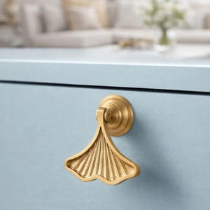 May include: A gold-colored, ginkgo leaf-shaped drawer pull is mounted on a light blue cabinet. The drawer pull has a detailed, fan-like design and a circular base. The background is blurred, showing a glimpse of a living room.