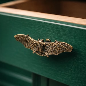 以下が含まれることがあります： 暗い緑色の引き出しに取り付けられた、ブロンズ色のコウモリ型の引き出し取っ手。 コウモリの翼は広げられ、体の細部が見えます。 引き出しは開いており、木製の内部が見えます。