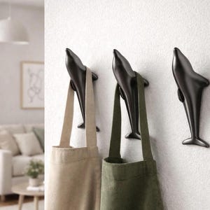 Puede incluir: Tres ganchos de pared negros con forma de delfín montados en una pared blanca. Dos bolsas de lona, una beige y otra verde oliva, cuelgan de los ganchos. Un elemento de decoración del hogar decorativo y funcional.