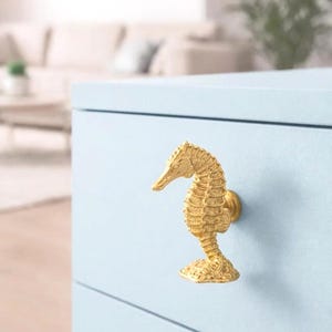 Könnte beinhalten: Goldfarbener Schrankknauf in Form eines Seepferdchens, montiert auf einer hellblauen Schublade. Der Knauf hat eine detaillierte, strukturierte Oberfläche und einen kleinen, runden Sockel. Der Hintergrund ist ein verschwommenes Interieur.