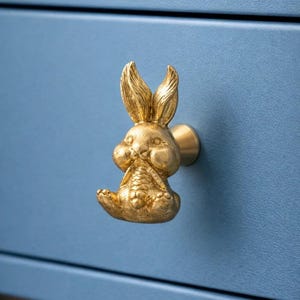 May include: A gold-colored bunny-shaped drawer pull mounted on a blue cabinet. The bunny is sitting with its legs tucked in and has detailed features. The cabinet has a textured surface.