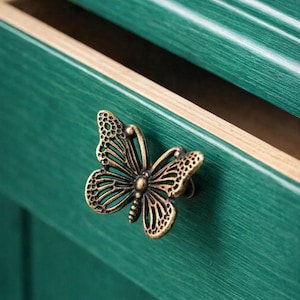 Può includere: Pomello per cassetto a forma di farfalla in bronzo su un mobile verde acqua. Il cassetto è parzialmente aperto, rivelando un interno marrone chiaro. Il pomello a farfalla presenta motivi dettagliati, aggiungendo un tocco decorativo al mobile.