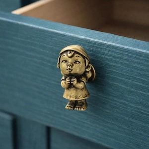 Peut inclure: Un bouton de meuble en laiton en forme de petite fille, avec un chapeau et une robe. Le bouton est fixé à un tiroir de meuble de couleur bleu sarcelle. Le tiroir est ouvert, révélant un intérieur marron clair.