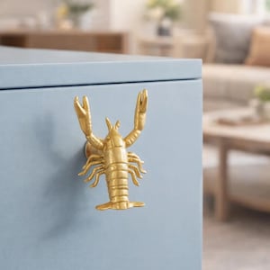 Peut inclure: Une poignée de meuble dorée en forme de homard est fixée sur une armoire bleu clair. La poignée de homard présente des pinces, des pattes et des segments de corps détaillés. L'armoire fait partie d'un meuble plus grand.