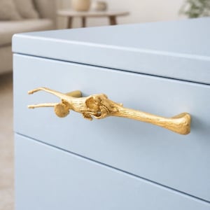 Può includere: Una maniglia dorata a forma di ramo è montata su un mobiletto azzurro. L'esclusiva maniglia aggiunge un tocco di design ispirato alla natura al mobile. Il mobile ha tre cassetti.