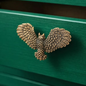 May include: A bronze-colored owl-shaped cabinet pull with outstretched wings. The pull is mounted on a green painted wooden drawer. The owl's feathers are detailed, and the hardware adds a decorative touch to the furniture.