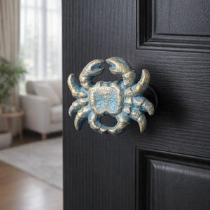 Op de afbeelding: Decoratieve deurknop in de vorm van een krab, beschilderd in blauwe en gouden tinten, bevestigd op een zwarte houten deur. De deur heeft een paneelontwerp. De achtergrond toont een woonkamer met een bank en een raam.