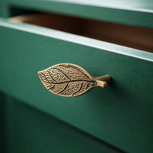 Peut inclure: Gros plan d'une poignée de meuble dorée en forme de feuille sur une armoire vert foncé. La poignée de feuille présente des nervures détaillées et une surface texturée. L'armoire est peinte d'un vert profond et riche.