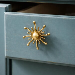 May include: A gold-colored sunburst drawer pull is mounted on a blue-gray painted wooden drawer. The drawer is partially open, revealing the interior. The sunburst design features a central sphere with radiating arms ending in small spheres.