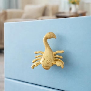 May include: A gold-colored scorpion-shaped cabinet knob mounted on a light blue drawer. The scorpion has a curved tail and outstretched claws. The knob adds a decorative touch to furniture.