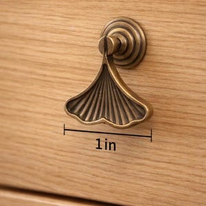 May include: A bronze-colored cabinet pull with a ginkgo leaf design, measuring 1 inch across. The pull is mounted on a light-colored wooden drawer, showcasing the hardware's intricate details and warm tones.