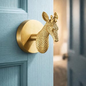 Puede incluir: Un tirador de puerta dorado con forma de cabeza de jirafa montado en una puerta azul claro. El tirador tiene una base circular y un diseño detallado de cabeza de jirafa. La puerta es de madera.
