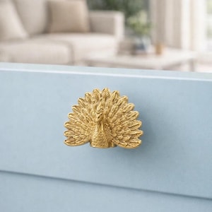 May include: A gold peacock drawer pull is fixed to a pale blue surface. The decorative handle features a detailed peacock design with fanned tail feathers. The background is blurred, suggesting a room setting. The handle is likely for a drawer or cabinet.
