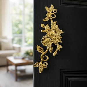 Op de afbeelding: Een goudkleurige bloemen deurgreep op een zwarte deur. De greep heeft een ontwerp van bloemen, bladeren en wijnranken. De achtergrond toont een woonkamer met een raam en meubels.