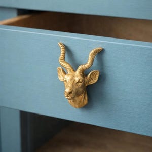 Op de afbeelding: Een goudkleurige kastknop in de vorm van een antilopekop is gemonteerd op een blauwe kastlade. De lade is open en onthult een houten interieur. De knop heeft gedetailleerde hoorns en gelaatstrekken.