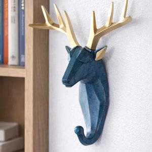 以下が含まれることがあります： 青と金の鹿の頭の壁掛けフック。鹿の頭は幾何学的なデザインで、下部にフックが付いています。角は金色で、頭は青色です。フックは白い壁に取り付けられています。
